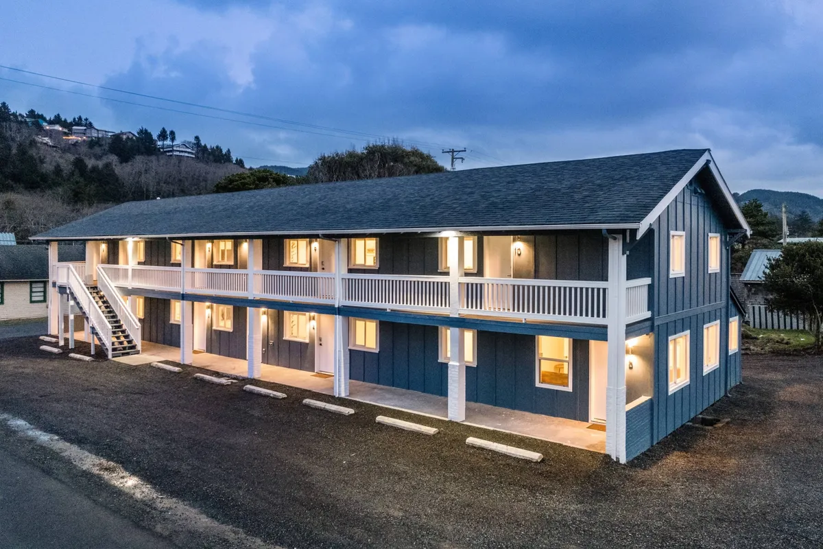 The building’s illuminated facade glows in the evening, with windows exuding warmth and a staircase inviting guests up. The balcony hints at community and outdoor enjoyment, while the setting evokes a welcoming and cozy mood.
