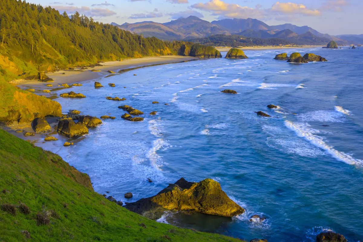 A scenic view of gentle ocean waves rolling onto the sandy beach, with iconic rock formations and mountains visible in the distance. The image invites thoughts of beachcombing, relaxing by the shore, and soaking in the beautiful coastal setting.