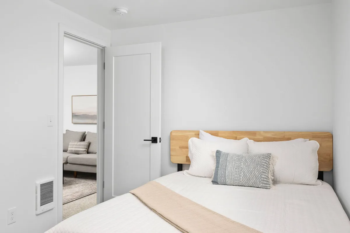 A cozy bedroom featuring a neatly made bed with decorative pillows, gentle natural light streaming in through the windows, and a doorway leading to another part of the unit. The ambiance is serene and comfortable, offering a glimpse of the welcoming interiors.