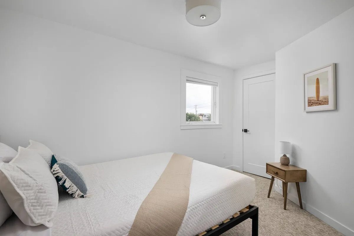 A serene interior bedroom with a neatly made bed dressed in crisp linens and decorative pillows. A wooden nightstand sits next to the bed, topped with a reading lamp, and a window allows in soft natural light, creating a tranquil atmosphere perfect for relaxation.